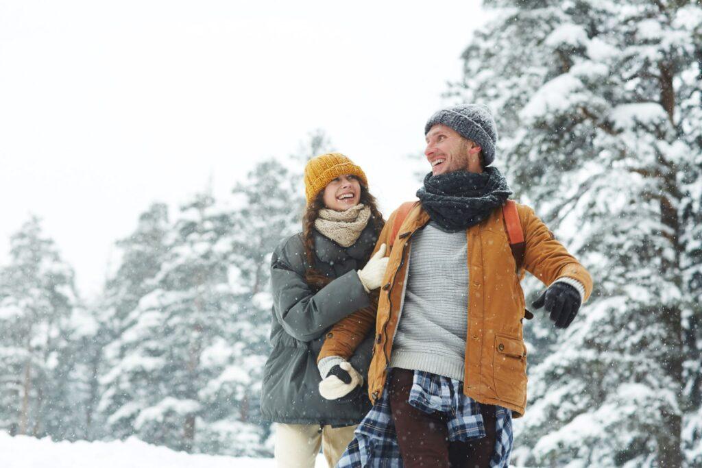 Ein lachendes Paar in Winterkleidung genießt einen verschneiten Tag im Freien, umgeben von schneebedeckten Bäumen. Der Winter bietet ideale Bedingungen für Aktivitäten wie Schlittschuhlaufen mit CCM Schlittschuhen.