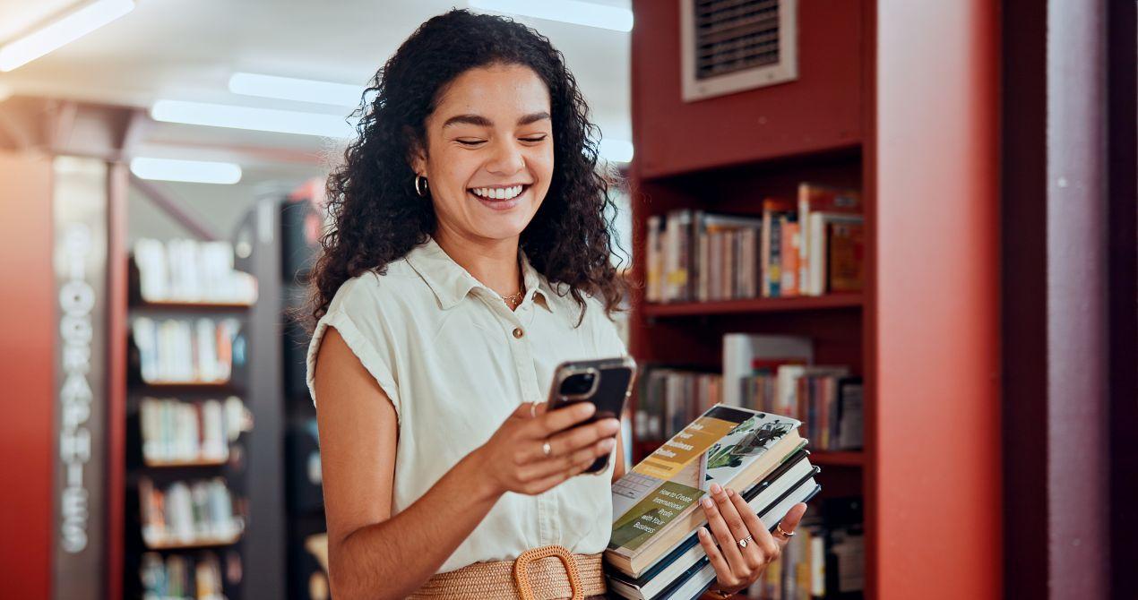 Studentin in Bibliothek mit Buechern und Smartphone als Inspiration fuer Bachelorarbeiten schreiben lassen